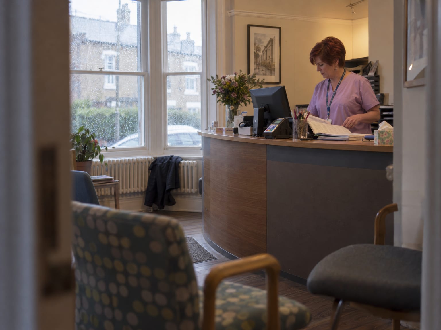 A dental receptionist at the front desk turning the page of an appointment book, rain on the window