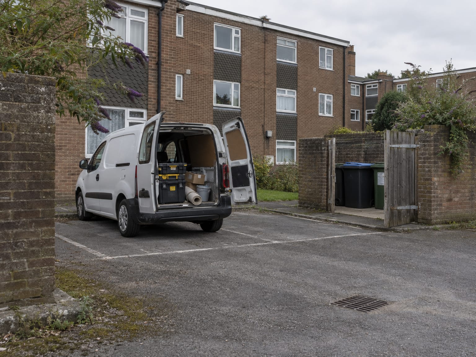 A small white van with its rear doors open in the car park behind a block of flats