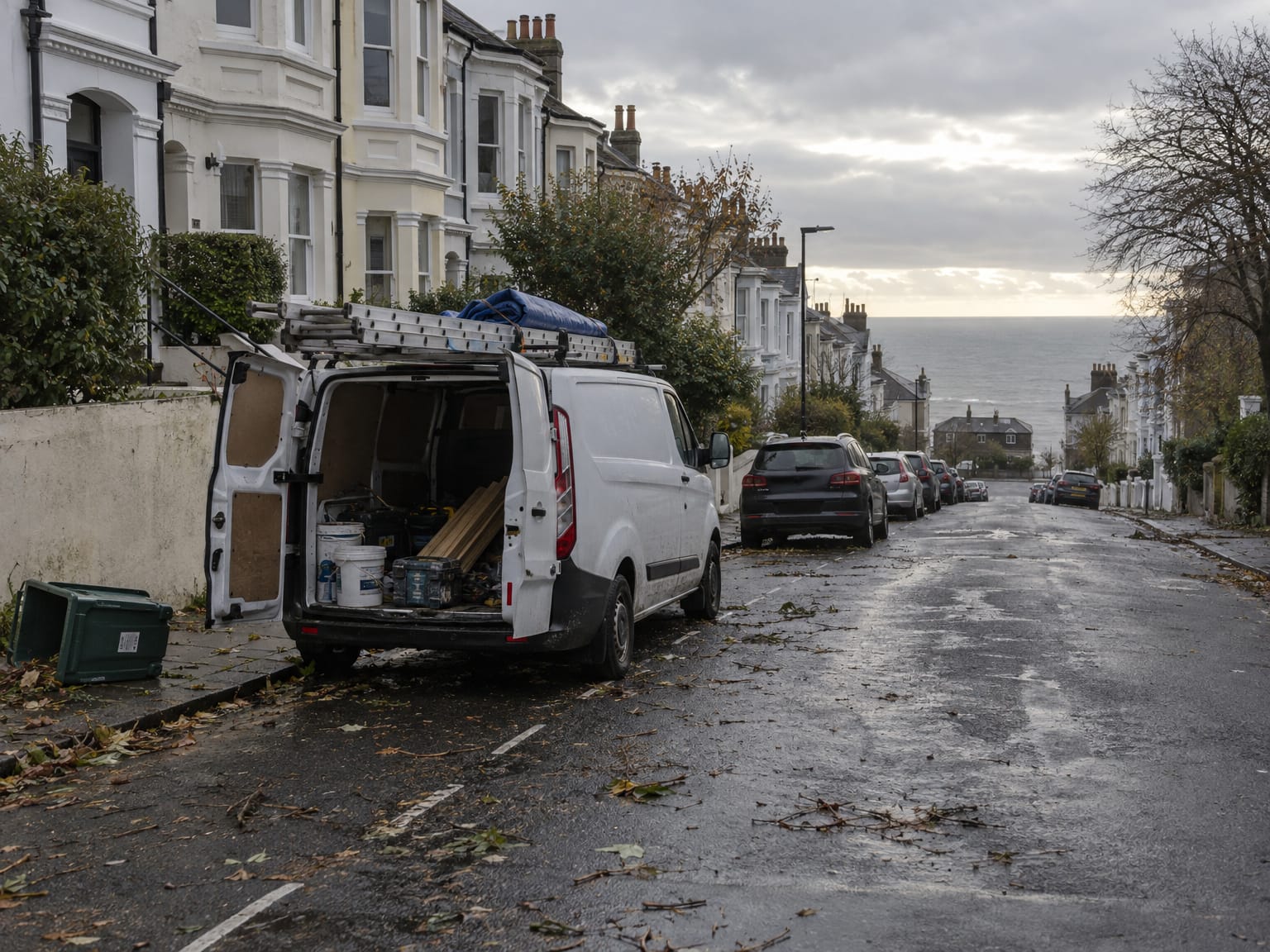 A white van with a ladder on the roof rack on a steep terraced street, grey sea at the bottom
